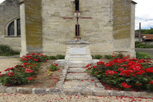 Ein steinernes Kreuz steht vor einer Kirche mit roten Rosen im Vordergrund, umgeben von Pflanzen und Bäumen, unter einem bewölkten Himmel.