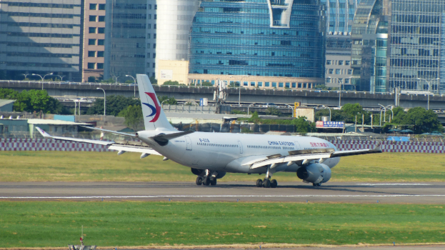 Großes Passagierflugzeug beim Start von einer Flughafenpiste mit umgebenem Gras, Zaun, Bäumen, Straßenlaternen, Schildern, Brücke, Gebäuden und bewölktem Himmel.