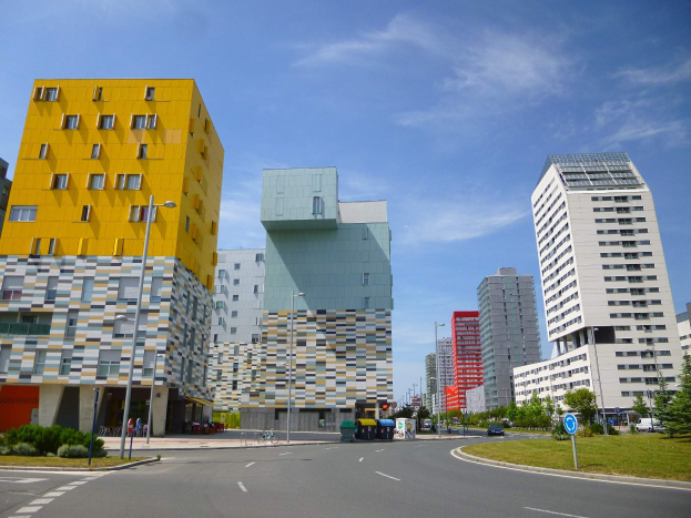 Eine Straße in der Stadt mit hohen Gebäuden, die als Teil der Universität Rotterdam gekennzeichnet sind, Straßenlaternen, Straßenbeleuchtung, Kraftfahrzeuge, Informationsschilder, Büsche, Bäume und ein bewölkter Himmel.